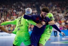 Two handball players compete intensely for the ball during a match, with one in a blue jersey holding the ball while the other in a neon green jersey attempts to block or tackle.