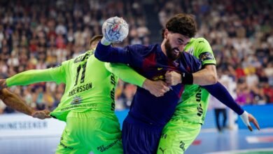 Two handball players compete intensely for the ball during a match, with one in a blue jersey holding the ball while the other in a neon green jersey attempts to block or tackle.